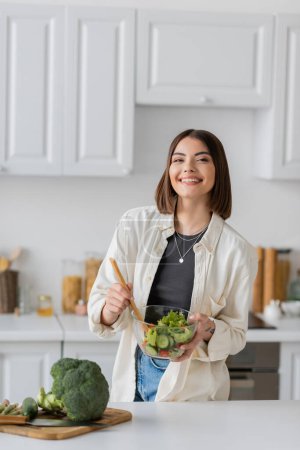 Positive Frau mischt frischen und vegetarischen Salat in Küche 