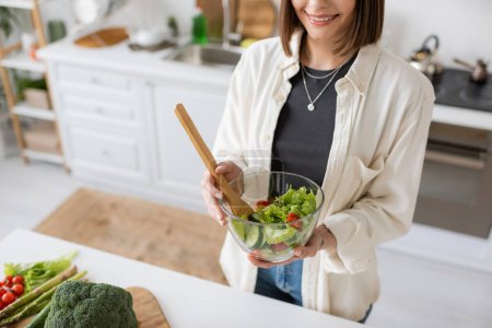 Vista recortada de una mujer sonriente sosteniendo ensalada fresca en un tazón en la cocina 