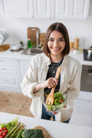Mujer joven positiva sosteniendo ensalada fresca y mirando a la cámara en la cocina 