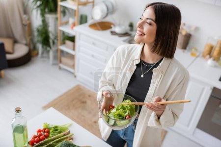 Carefree woman holding fresh salad near vegetables and olive oil in kitchen 