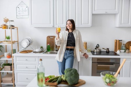 Mujer morena sonriente sosteniendo jugo de naranja cerca de ensalada borrosa en la cocina 