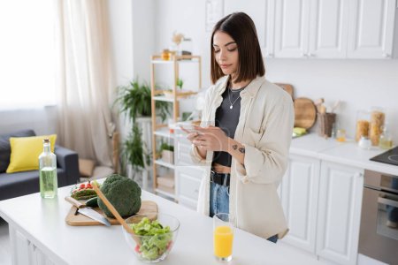 Tattooed woman using smartphone near orange juice and fresh salad in kitchen 