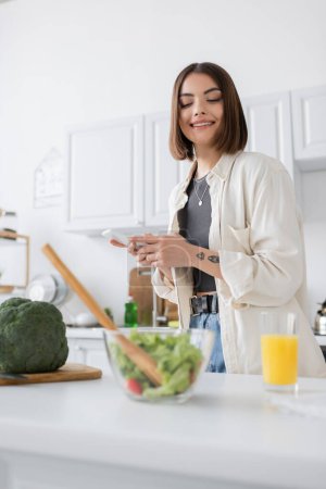 Tätowierte Frau mit Smartphone in der Nähe von Salat und Orangensaft in der Küche 