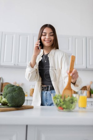 Smiling woman talking on smartphone and mixing fresh salad in kitchen 