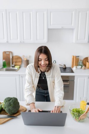 Lächelnde Frau mit Laptop neben frischem Salat und Orangensaft in der Küche 