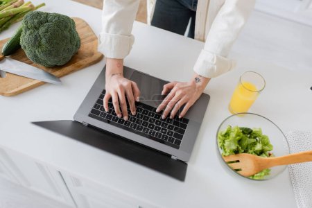 Cropped view of tattooed freelancer using laptop near salad and vegetables in kitchen 