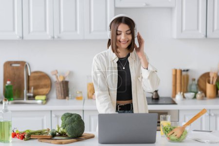 Cheerful young woman in headphones using laptop near salad and fresh vegetables in kitchen 