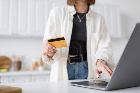 Ausgeschnittene Ansicht einer verschwommenen Frau mit Kreditkarte bei der Bestellung von Lebensmitteln auf dem Laptop zu Hause 