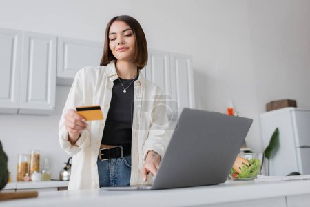 Brunette woman holding credit card near laptop and fresh salad in kitchen 