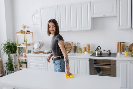 Joven mujer tatuada limpiando encimera con trapo en la cocina en casa 
