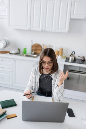 Téléchargez les photos : Freelance dans les lunettes à l'aide d'un ordinateur portable près des ordinateurs portables et carte de crédit dans la cuisine - en image libre de droit