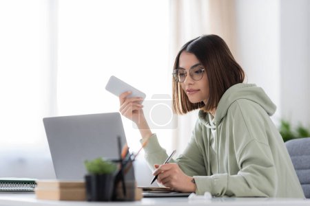 Brunette freelancer in eyeglasses holding cellphone and writing on notebook near laptop at home 