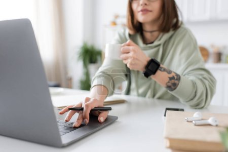 Vista recortada del freelancer borroso sosteniendo la taza de café y usando el ordenador portátil cerca del libro y los auriculares en casa 