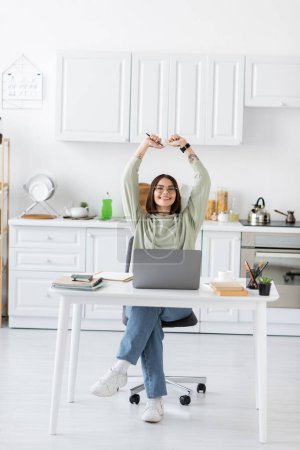 Téléchargez les photos : Indépendante excitée dans les lunettes montrant un geste oui près d'un ordinateur portable et des ordinateurs portables dans la cuisine - en image libre de droit