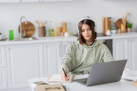Téléchargez les photos : Jeune pigiste dans un casque d'écriture sur ordinateur portable près de gadgets et café sur la table à la maison - en image libre de droit