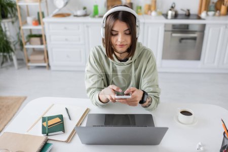 Téléchargez les photos : Brunette freelance dans les écouteurs en utilisant smartphone près d'un ordinateur portable et café à la maison - en image libre de droit