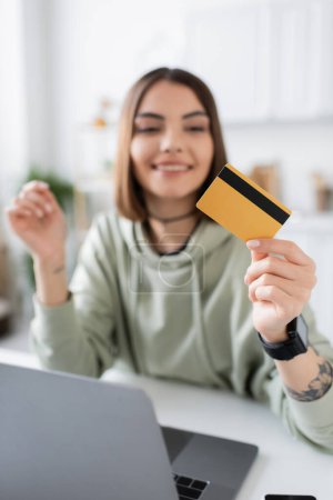 Téléchargez les photos : Femme brune joyeuse tenant une carte de crédit près d'un ordinateur portable pendant les achats en ligne à la maison - en image libre de droit