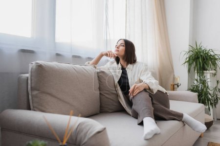 Junge Frau in lässiger Kleidung schaut weg, während sie zu Hause auf der Couch sitzt 