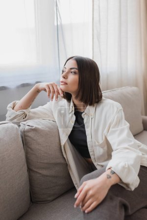 Young tattooed woman looking away while sitting on couch at home 
