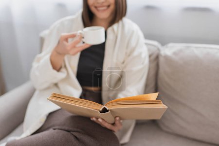 Vue recadrée d'une femme floue tenant une tasse de café et un livre de lecture sur le canapé à la maison 