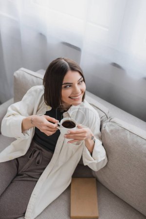 Hochwinkelblick der fröhlichen brünetten Frau, die eine Tasse Kaffee in der Nähe von Buch auf der Couch im Wohnzimmer hält 