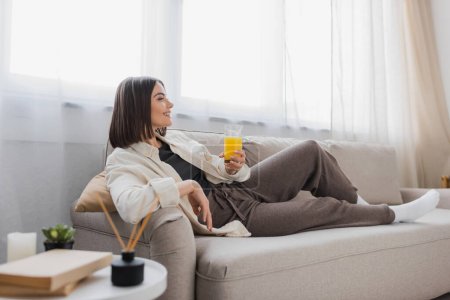 Téléchargez les photos : Vue latérale d'une femme brune souriante tenant un verre de jus d'orange alors qu'elle était assise sur le canapé à la maison - en image libre de droit