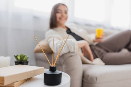 Aroma sticks near books and blurred woman sitting on couch at home 