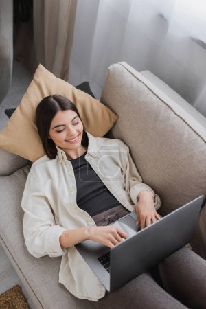 Foto de Vista superior de la mujer morena alegre usando el ordenador portátil mientras está acostado en el sofá en casa - Imagen libre de derechos