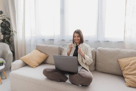 Excited freelancer showing yes gesture and looking at laptop on couch at home