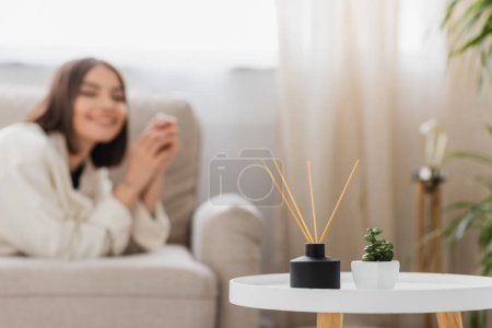 Planta y bambú palos perfumados en la mesa de café cerca de la mujer borrosa en el sofá en casa 