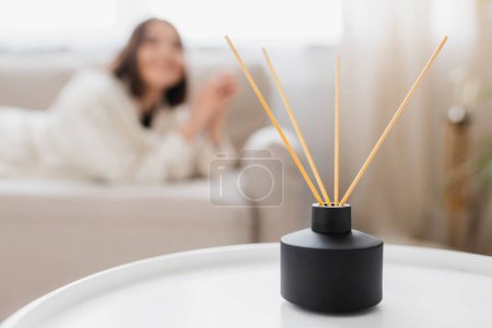 Bamboo sticks in aroma diffuser on coffee table near blurred woman in living room 