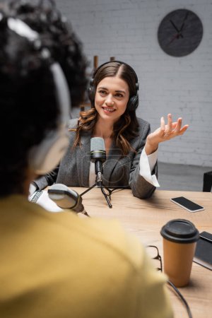 charming brunette interviewer in headphones and grey blazer talking to blurred indian man near microphones, coffee to go, smartphone and notebook on table in broadcasting studio