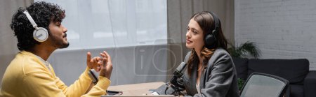 Photo for Side view of young and curly indian man in headphones and yellow jumper talking and gesturing near microphones and smiling brunette interviewer wearing grey blazer in radio studio, banner - Royalty Free Image