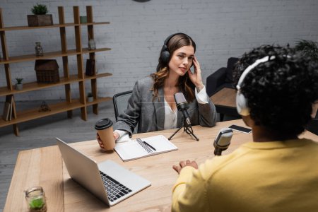 Photo for Tattooed brunette interviewer in headphones holding takeaway drink and looking at blurred indian man near laptop, microphones, notebook and mobile phone on table in broadcasting studio - Royalty Free Image