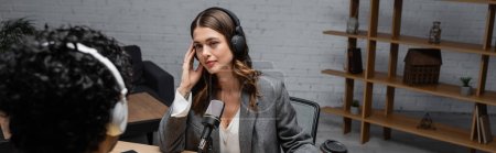 Photo for Brunette radio host in grey blazer adjusting headphones during interview with blurred indian man near microphone and paper cup with wooden rack on background in broadcasting studio, banner - Royalty Free Image