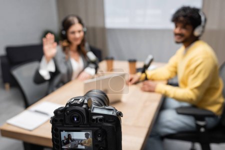 Téléchargez les photos : Caméra numérique professionnelle enregistrement interview de femme agitant la main et jeune invité indien assis à la table avec ordinateur portable et tasses en papier floues dans le studio de radiodiffusion - en image libre de droit