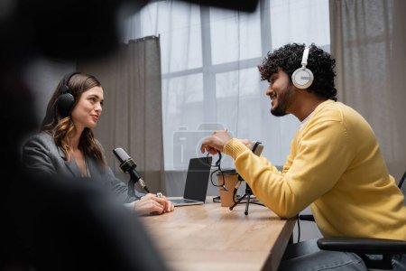 Photo for Side view of smiling and curly indian man in headphones and yellow jumper talking to brunette interviewer near laptop with blank screen and coffee to go in radio studio on blurred foreground - Royalty Free Image