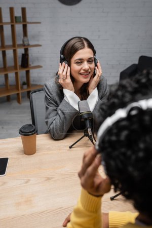 happy brunette broadcaster adjusting headphones during interview with blurred indian man near professional microphone, coffee to go and smartphone with blank screen in modern radio studio