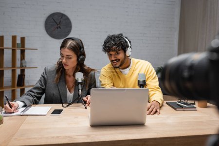 Photo for Brunette radio host in headphones writing in notebook close to positive indian colleague looking at laptop near professional microphones and smartphone with blank screen in studio - Royalty Free Image