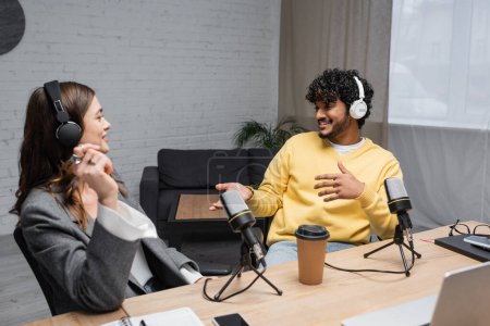 happy multiethnic radio hosts in headphones talking near professional microphones, coffee to go, notebooks and smartphones on table in modern broadcasting studio with armchair on background