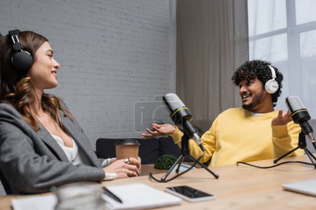 joyful indian man in headphones and yellow jumper gesturing and talking to smiling colleague sitting with paper cup near microphones and blurred notebook with smartphone in broadcasting studio