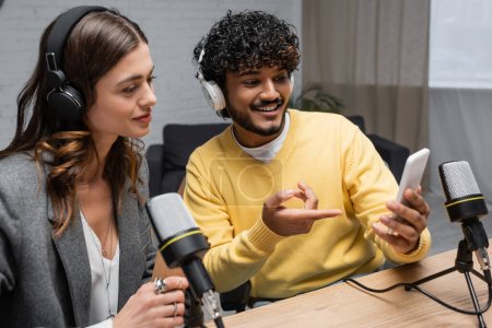 smiling indian podcaster in headphones and yellow jumper showing mobile phone to charming brunette colleague near professional microphones in broadcasting studio