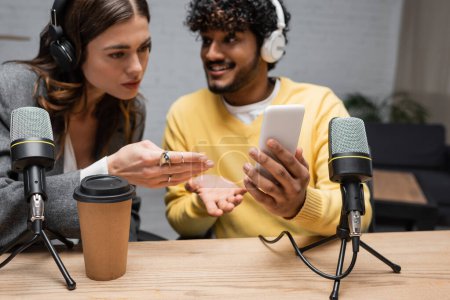 serious radio host in headphones pointing at mobile phone in hands of positive indian colleague in yellow jumper near coffee to go and microphones in broadcasting studio
