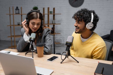 brunette podcaster in headphones talking in microphone close to young and happy indian colleague in yellow jumper near laptop, notebooks and smartphone with blank screen in studio