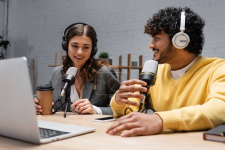 souriant radiodiffuseurs interraciaux dans les écouteurs travaillant avec des microphones professionnels près d'un ordinateur portable, café à emporter et téléphone portable avec écran blanc tout en enregistrant podcast dans un studio de radio