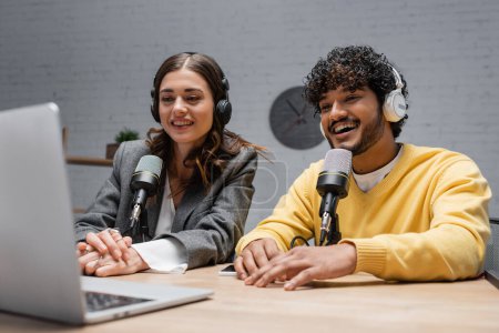 fröhliche multiethnische Radiomoderatoren mit Kopfhörern, gelbem Pullover und grauer Jacke, die auf Laptop schauen, während sie in professionellen Mikrofonen sprechen und im modernen Studio einen Podcast aufnehmen 