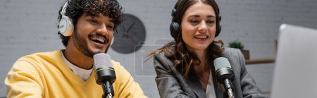 Foto de Hombre indio excitado y rizado en jersey amarillo con encantadora mujer morena en chaqueta gris hablando en micrófonos profesionales mientras trabajaba y grababa podcast en el estudio de radio, pancarta - Imagen libre de derechos