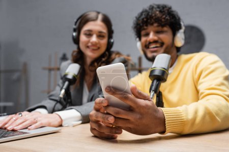 cheerful indian radio host in headphones and yellow blazer holding smartphone near brunette colleague using laptop close to professional microphones in radio studio