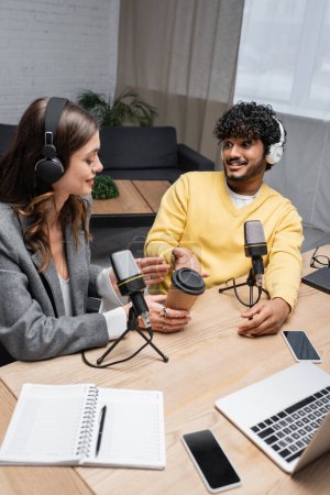 Foto de Hombre indio alegre en auriculares y mujer morena apuntando a la taza de papel cerca de micrófonos profesionales, portátil, portátiles y teléfonos inteligentes con pantalla en blanco en el estudio de radio - Imagen libre de derechos