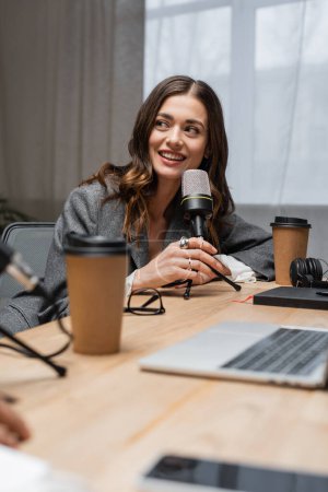 happy and charming radio host looking away while talking in professional microphone near paper cups, blurred laptop and notebook on table in modern photo studio